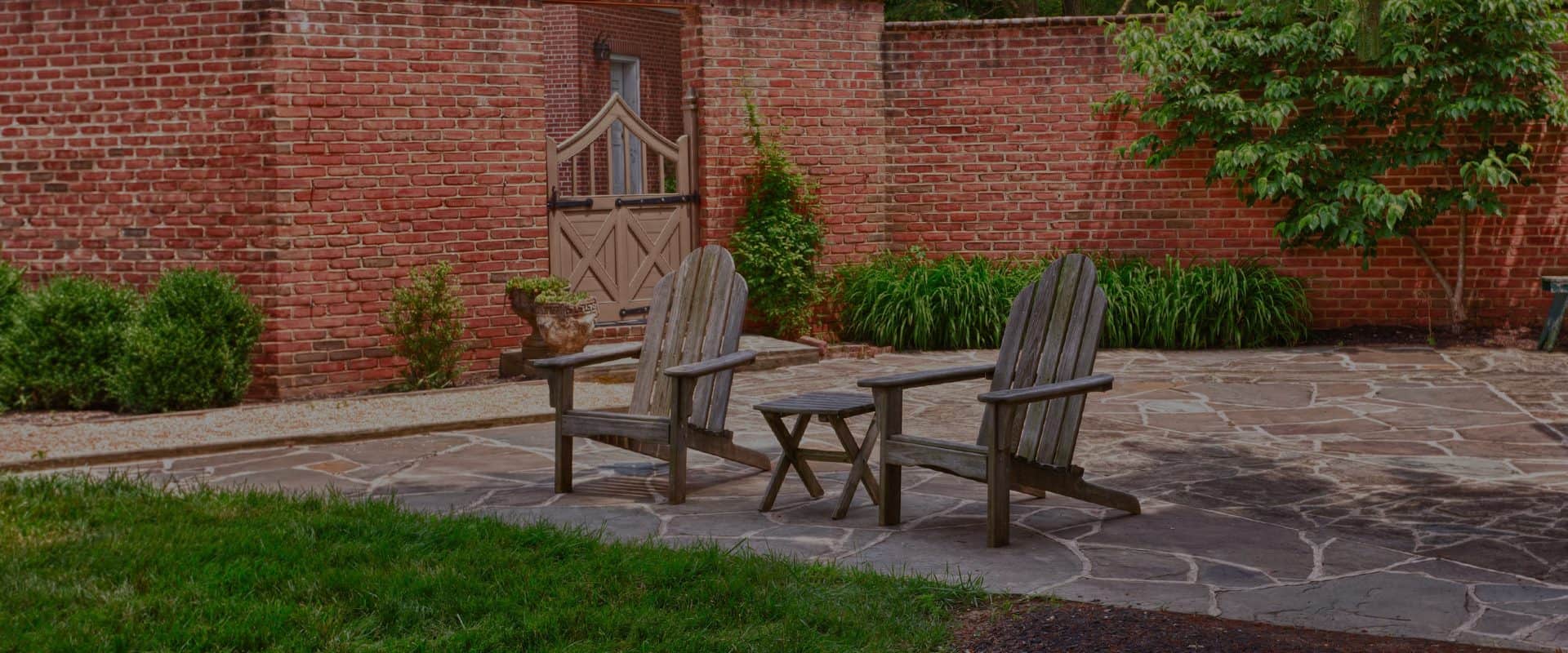 A cozy outdoor courtyard with two wooden Adirondack chairs and a small table on a stone patio by Danbury Landscaping. The area is enclosed by a brick wall with a wooden door, and greenery, including bushes and a tree, adds a touch of nature.