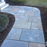 A curved stone walkway with large rectangular tiles of various shades of gray and tan leads to a set of stairs. The path, expertly crafted by roots landscaping ct, is bordered by dark soil and small, neat bushes on the left side. The surrounding grass is partially visible.