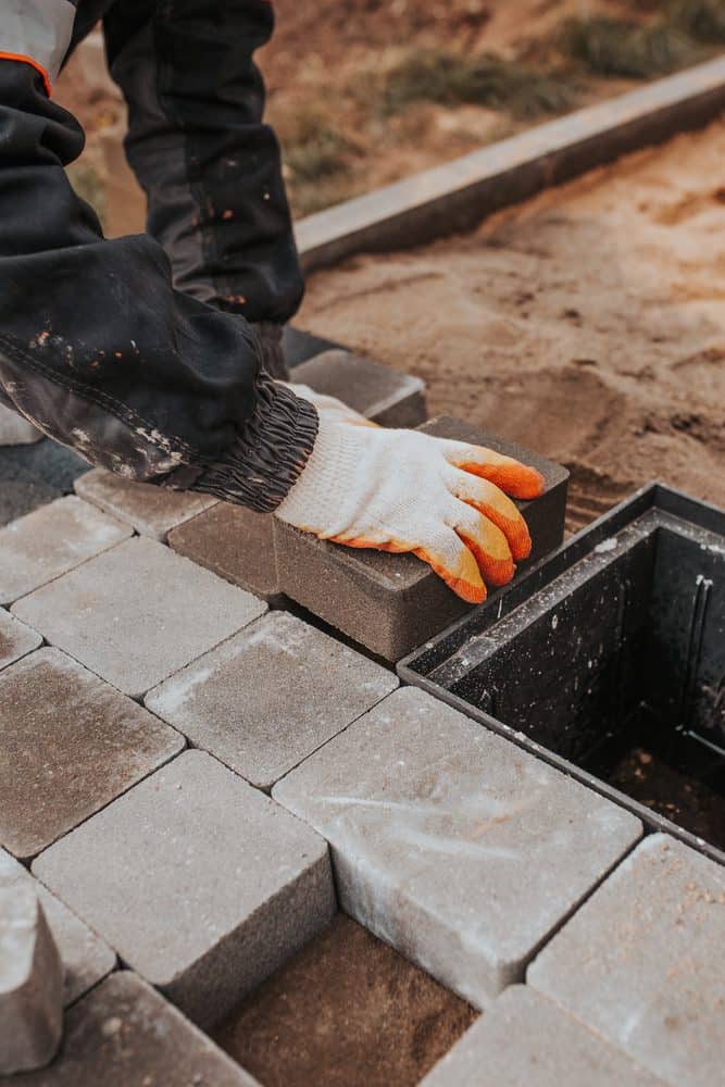 A person wearing work gloves is placing a rectangular stone block on a patch of laid stones. The surroundings include sand and other stone blocks, indicating a paving or construction project in progress, typical of masonry services Connecticut.