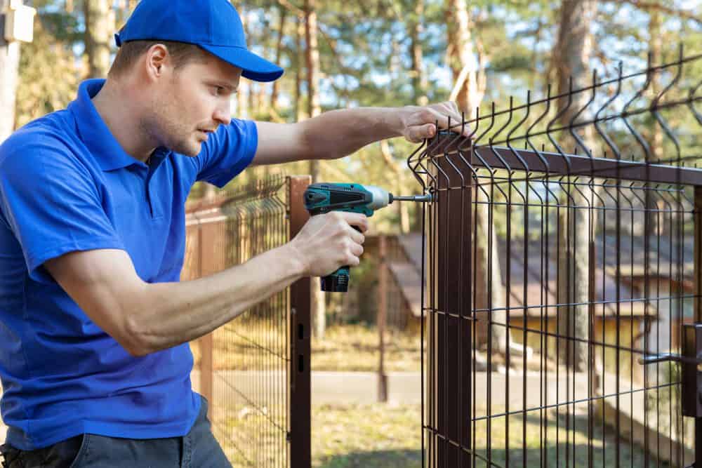 A man wearing a blue polo shirt and cap uses a cordless drill to secure a wire fence to a brown metal post in an outdoor setting with trees in the background, showcasing the precision and expertise of Fencing Services Connecticut.