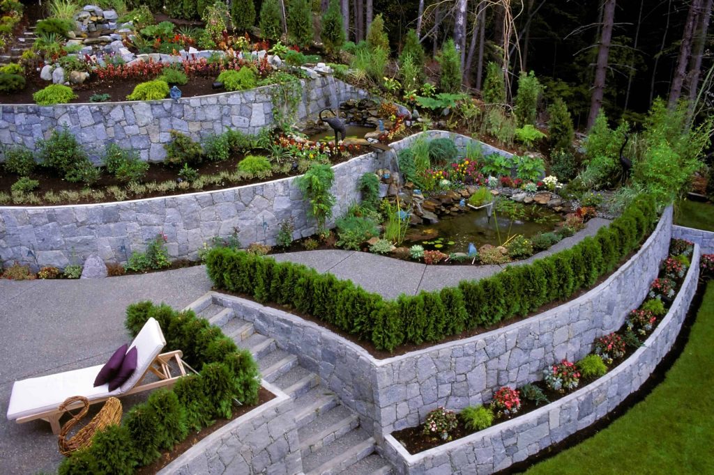 A beautifully landscaped multi-tiered garden with stone walls crafted by expert masonry services in Connecticut features a small pond with a fountain and lush greenery. Flower beds with colorful blooms, neatly trimmed shrubs, and a pathway leading to different levels complete this serene setting, where a lounge chair sits on one tier.