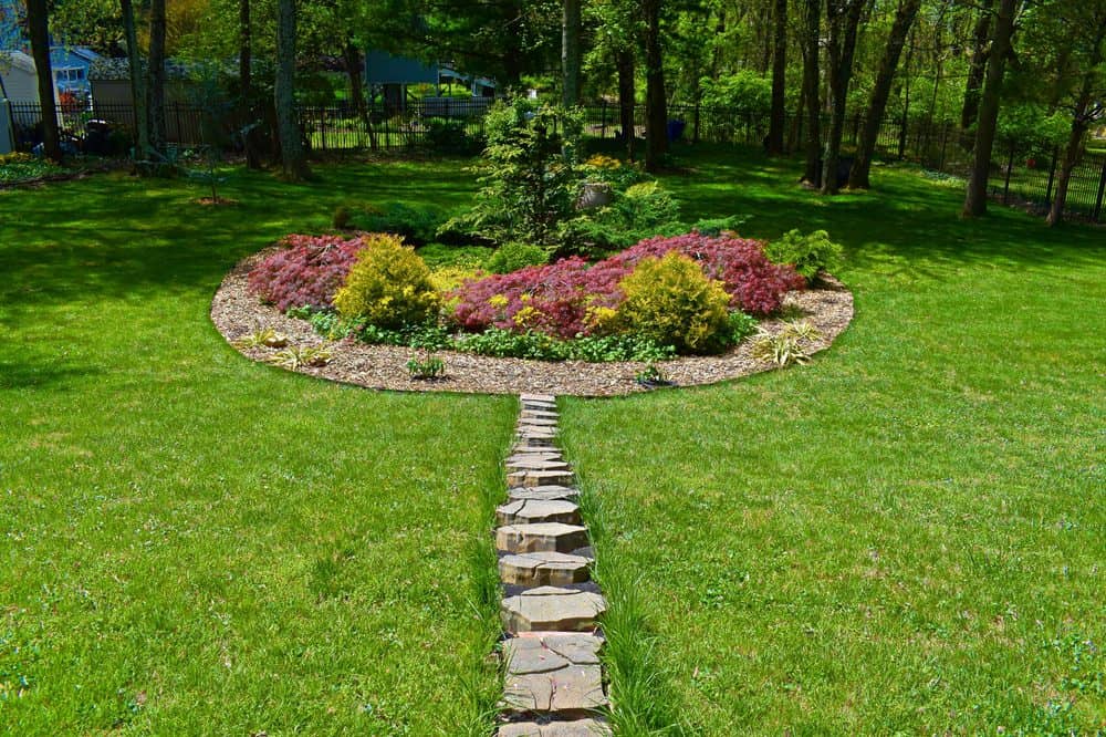 A lush garden features a circular flowerbed with colorful plants and shrubs, embraced by expert landscaping. A stone path leads directly to the flowerbed, and subtle masonry elements integrate seamlessly. Trees create a natural backdrop in the distance, providing tranquility.