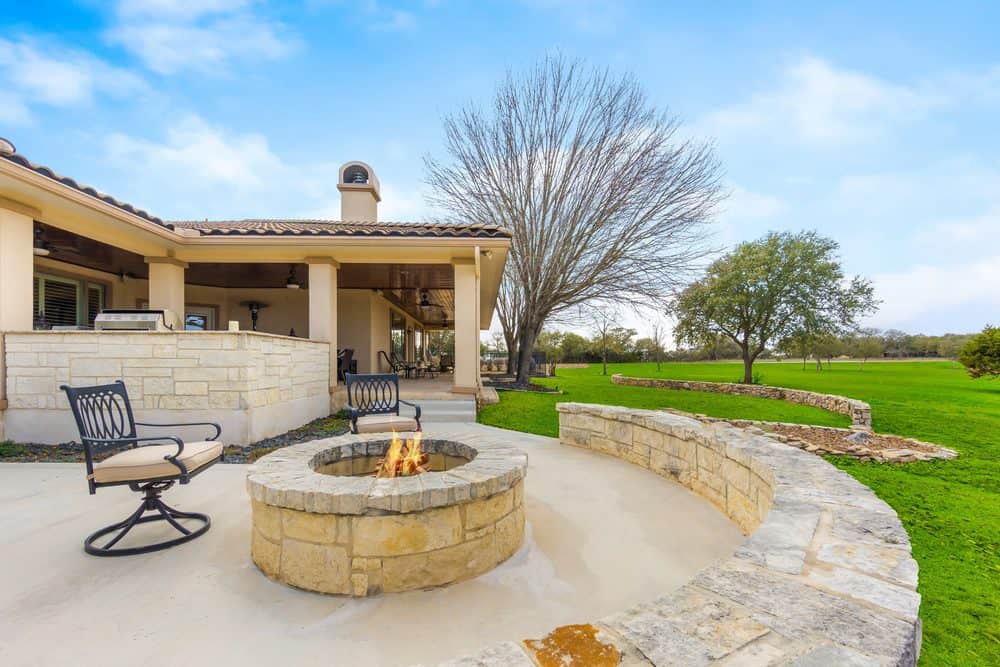 An inviting backyard with a stone fire pit surrounded by a curved stone seating area, crafted by expert Masonry Services Connecticut. To the left, there's a covered patio attached to the house, featuring a couple of chairs and an outdoor kitchen. The background includes a lush green lawn and a few trees.