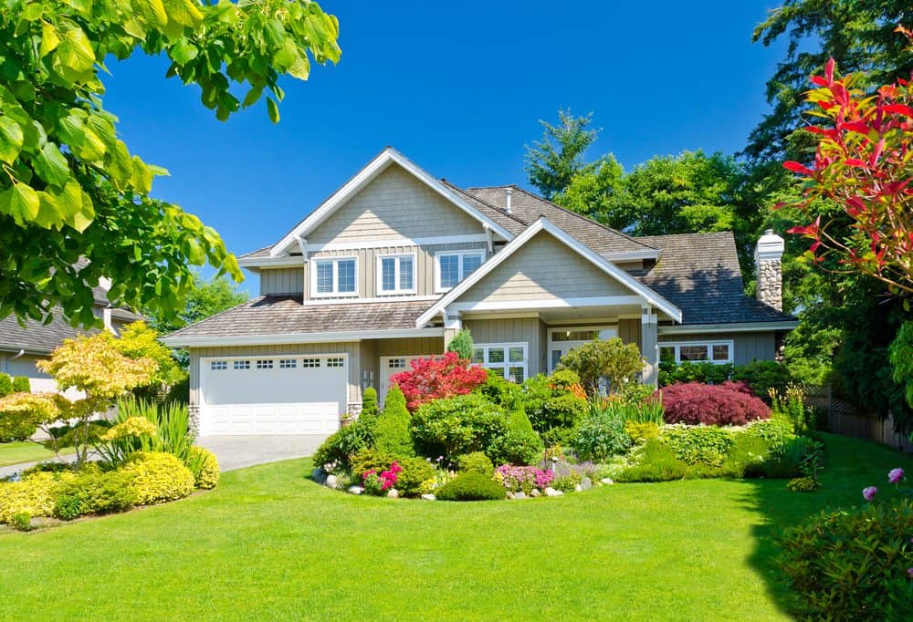 A two-story house with a neatly manicured lawn and colorful flower garden. The house has a gray exterior with white trim, a double garage, and large windows. It is surrounded by lush greenery, trees, and expertly crafted masonry services in Connecticut under a clear blue sky.
