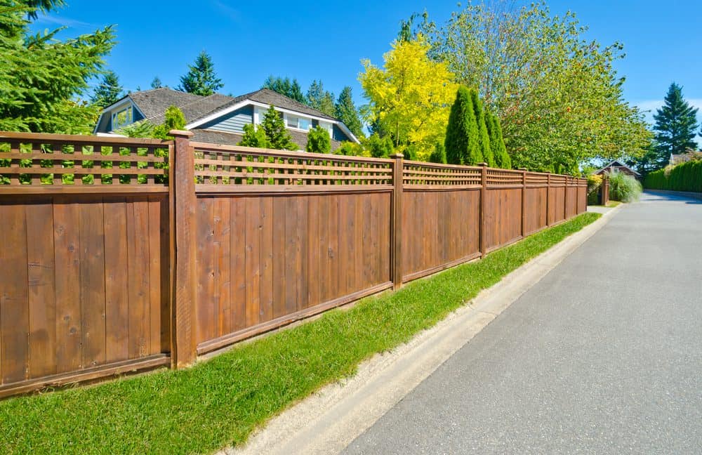 A well-maintained wooden fence borders a residential property on a sunny day. The fence, crafted by Fencing Services Connecticut, features a lattice design along the top and runs alongside a neatly trimmed green lawn, with a sidewalk and road adjacent to it. Trees and houses are visible in the background.