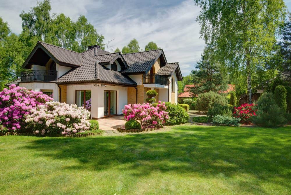 A modern two-story house with a dark shingled roof is surrounded by a well-maintained lawn and lush garden. Blooming pink and purple flowers line the walkway, while tall green trees provide a tranquil backdrop against a partly cloudy sky, showcasing superb Landscaping Services Connecticut.