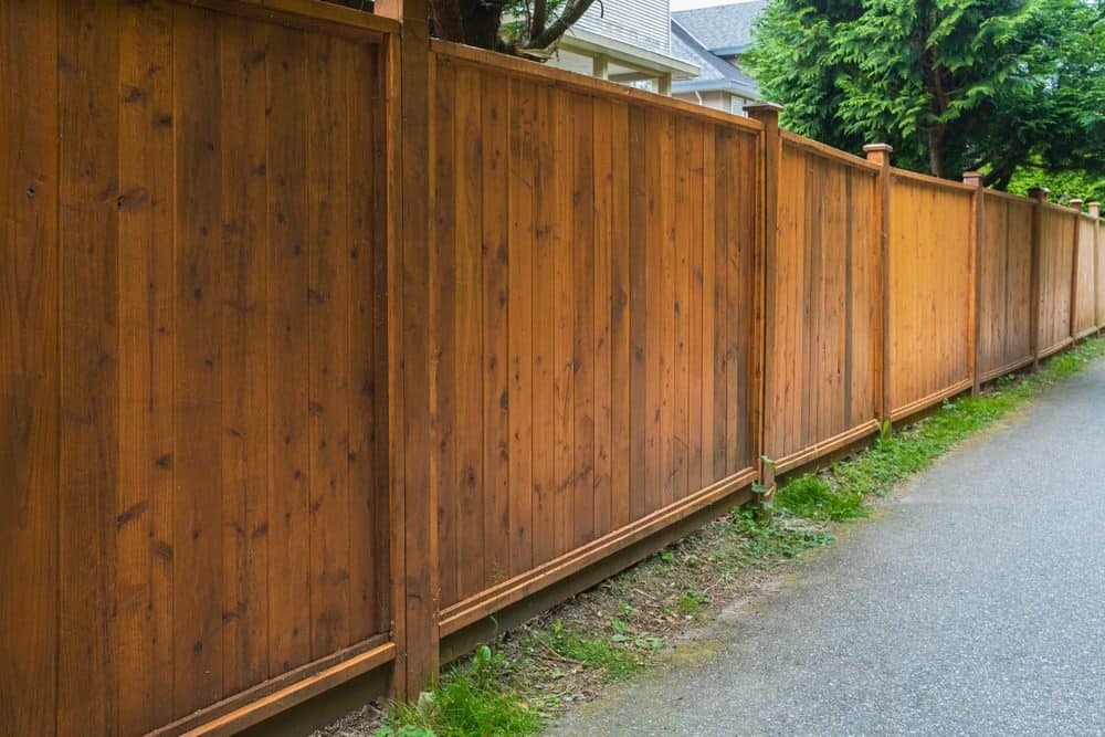 A long wooden privacy fence stretches alongside a paved pathway. The fence consists of vertical wooden planks framed by horizontal beams and supports, with a neatly kept grassy edge lining the bottom. Lush greenery and treetops are visible beyond the fence, courtesy of expert Landscaping Services Connecticut.