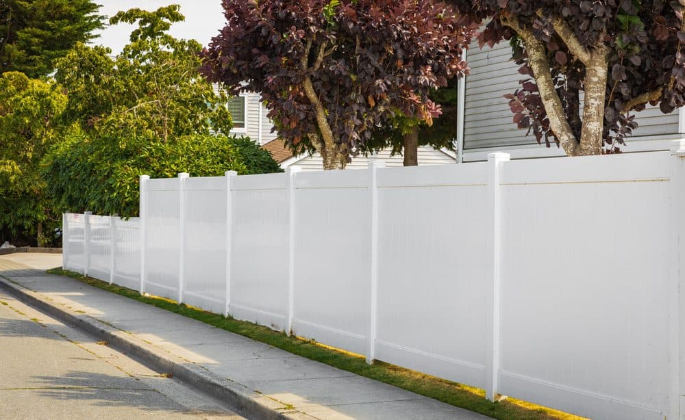 A long, white, vinyl privacy fence lines a sidewalk in a suburban neighborhood. The fence stands tall beside a neatly paved road, with lush green trees and well-maintained bushes in the background—courtesy of top-notch Landscaping Services Connecticut.