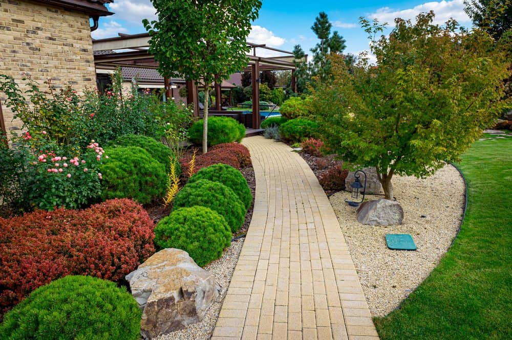 A well-maintained garden with a winding brick pathway, showcasing expert Landscaping Services Connecticut. Surrounding the path are neatly trimmed bushes, rocks, and a variety of trees and flowering plants. In the background, there is a modern wooden pergola and a house made of brick. The sky is partly cloudy.