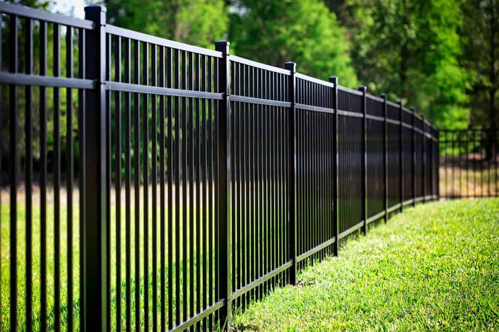 A black metal fence with vertical bars stands on a lush green lawn, expertly installed by Fencing Services Connecticut. Trees and greenery blur in the background, suggesting an inviting outdoor setting like a garden or yard. The sleek, modern fence stretches into the distance.