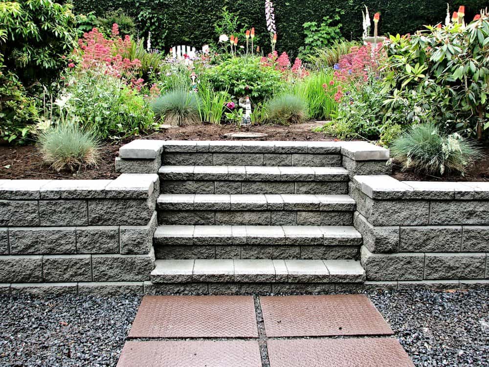 A stone staircase leads up through a landscaped garden, surrounded by lush greenery and colorful flowers. The steps are bordered by brick retaining walls, and the path is made of smooth, gray stones. The garden is vibrant and well-maintained, showcasing premium Landscaping Services Connecticut.