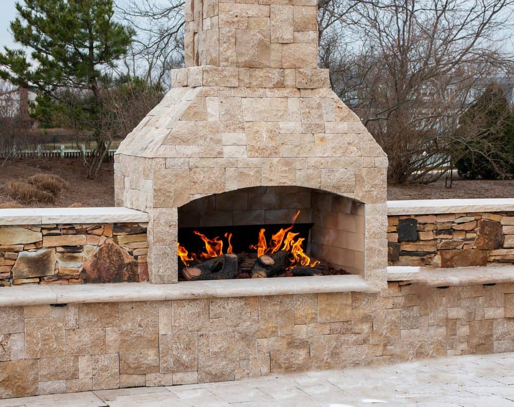 An outdoor stone fireplace with a burning fire, surrounded by a stone wall. The scene includes a paved ground and trees without leaves in the background, suggesting a colder season. Perfectly crafted through Masonry Services Connecticut, this setup brings warmth and charm to your outdoor space.