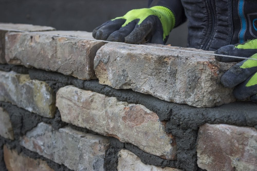A person wearing bright green gloves is constructing a brick wall. Carefully aligning and securing the bricks with mortar, they ensure a solid and even structure. The close-up highlights the rugged texture of the bricks and fresh, wet mortar, showcasing expert Masonry Services Connecticut can rely on.