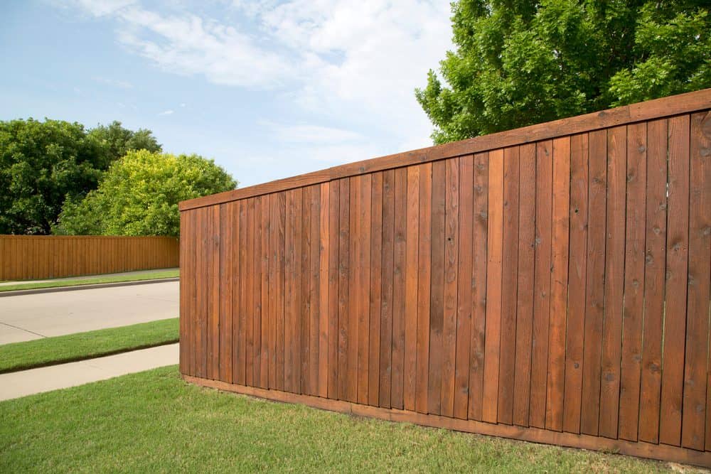A tall wooden fence runs parallel to a quiet suburban street, showcasing the expert craftsmanship of Fencing Services Connecticut. The fence is made of vertical wooden planks with a rich, reddish-brown stain. Green trees and a manicured lawn are visible on both sides of the street under a partly cloudy sky.