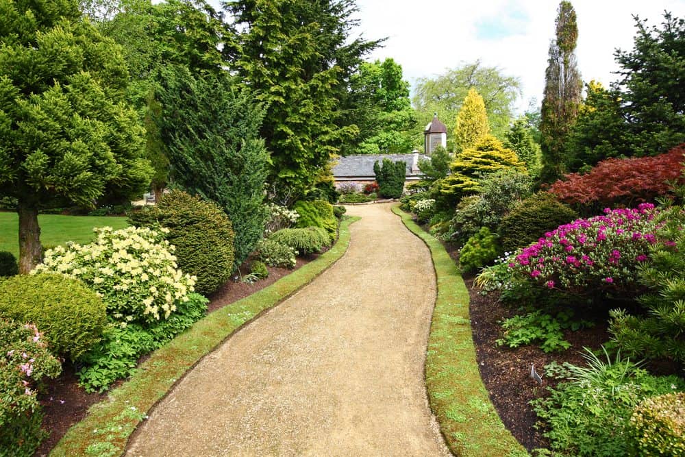 A serene garden path, bordered by lush greenery, shrubs, and colorful flowering plants, leads towards a quaint house with a small tower in the background. The pathway, masterfully crafted by Masonry Services Connecticut, is made of light-colored gravel, framed by well-manicured lawns and tall trees.