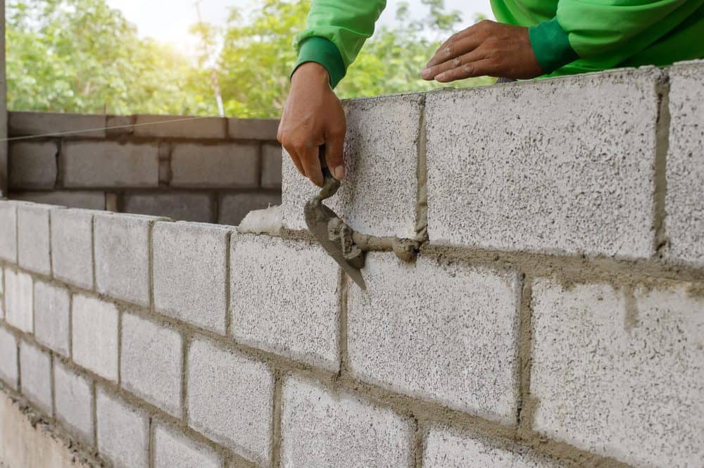 Person in a green shirt applying mortar with a trowel to cement blocks while building a wall. The background includes partially constructed cement walls and blurred greenery with sunlight filtering through, showcasing expert Masonry Services Connecticut.