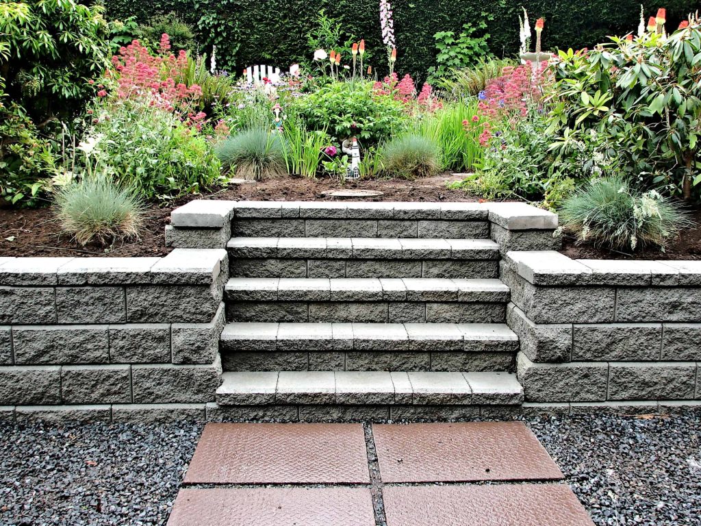 A garden landscape with stone stairs leads up to a flower bed. Rows of neatly arranged flower bushes in various colors surround the steps, and a lush green hedge forms the backdrop. The foreground has gravel and stepping stones, showcasing expert Landscaping Services Connecticut.