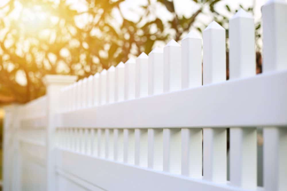 Close-up of a white picket fence with sunlight streaming through trees in the background, creating a warm and serene atmosphere. The fence features vertically aligned slats and a horizontal top bar, evoking a classic and neat aesthetic, perfect for enhancing your home—courtesy of Fencing Services Connecticut.