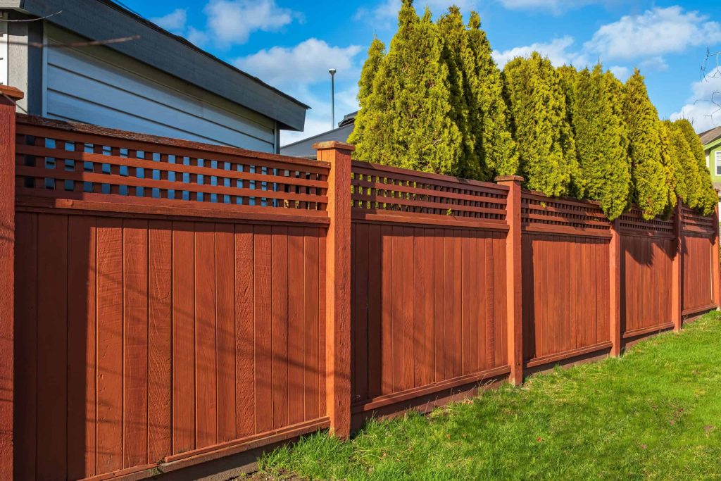 A tall, wooden privacy fence painted red-brown stands in a backyard, with decorative latticework at the top. Behind the fence, a row of green, bushy trees provides additional privacy. The scene is under a partly cloudy sky on a sunny day. Perfect for any yard needing Fencing Services Connecticut.