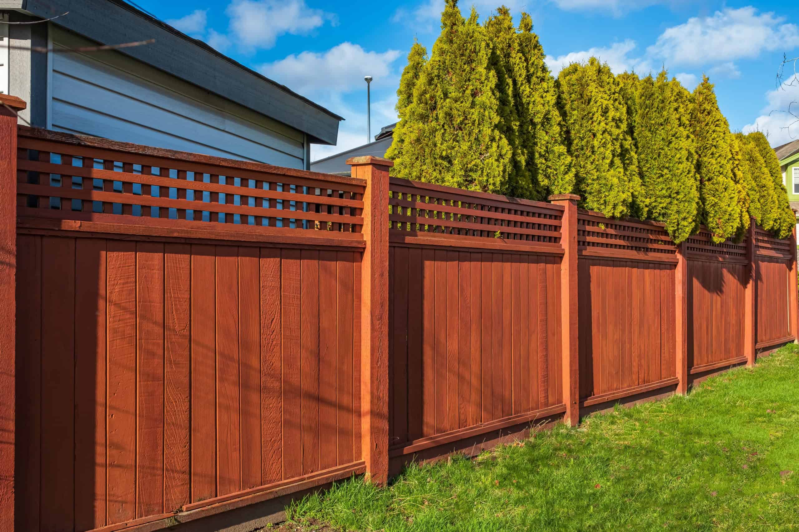 A tall, wooden privacy fence painted red-brown stands in a backyard, with decorative latticework at the top. Behind the fence, a row of green, bushy trees provides additional privacy. The scene is under a partly cloudy sky on a sunny day. Perfect for any yard needing Fencing Services Connecticut.