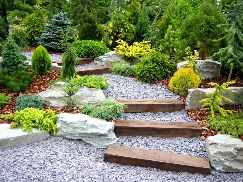 A serene garden with a stone pathway and wooden steps, surrounded by lush green shrubs, small plants, and decorative rocks. The natural greenery and varied textures create a tranquil, well-maintained landscape. Enhance your space further with expert Landscaping Services Connecticut.