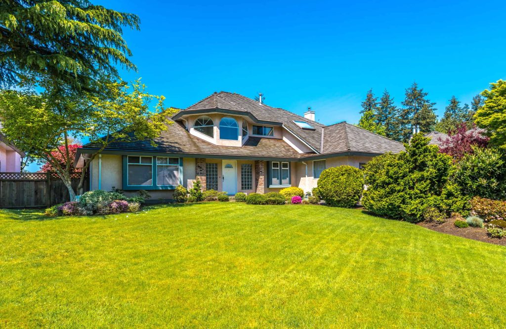 A two-story house with a steeply pitched roof and several windows is surrounded by a lush, well-maintained lawn courtesy of Landscaping Services Connecticut. Various shrubs and trees add to the charm. The sky is clear and blue, and the overall scene is bright and inviting.