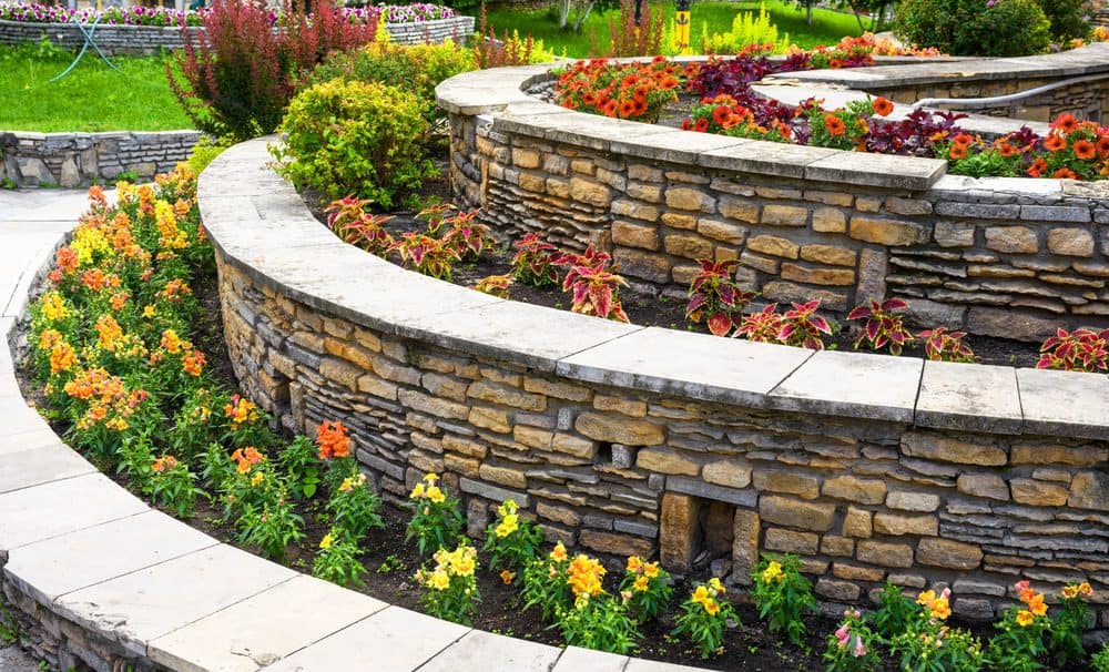 A beautiful terraced garden featuring various colorful flowers. The stone terraces, crafted by top-notch Masonry Services Connecticut, are lined with yellow, orange, and red blooms, creating a vibrant and lush landscape in a serene park setting. The curved stone walls add structure to the garden's design.