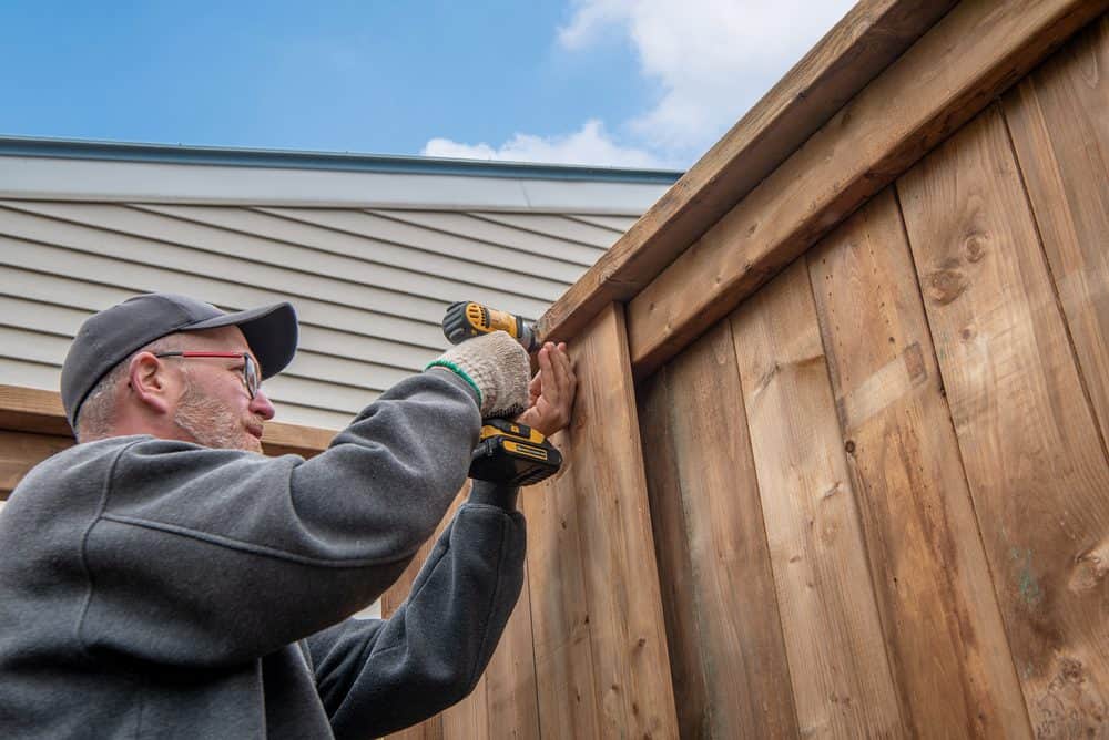 A person in a cap, glasses, and gloves is using a power drill to secure a piece of wood on a wooden fence. The setting appears to be outdoors near a building with a metal roof under the partly cloudy sky. This scene exemplifies the expert craftsmanship offered by Fencing Services Connecticut.