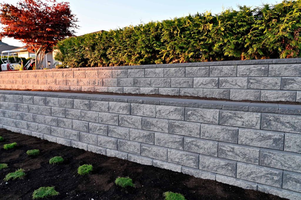 A landscaped garden featuring two tiers of gray stone retaining walls, courtesy of our Masonry Services Connecticut team, with lush green shrubs on the top tier. The ground in front of the walls is lined with small, freshly planted greenery. A tree with red foliage is visible on the left.