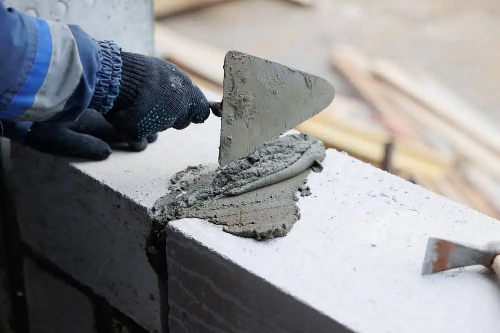 A person wearing black gloves and a blue jacket is using a trowel to apply mortar to the top of a concrete block wall, preparing it for the next layer of blocks. Construction materials and tools are visible in the blurred background, showcasing the quality of masonry services Connecticut residents rely on.