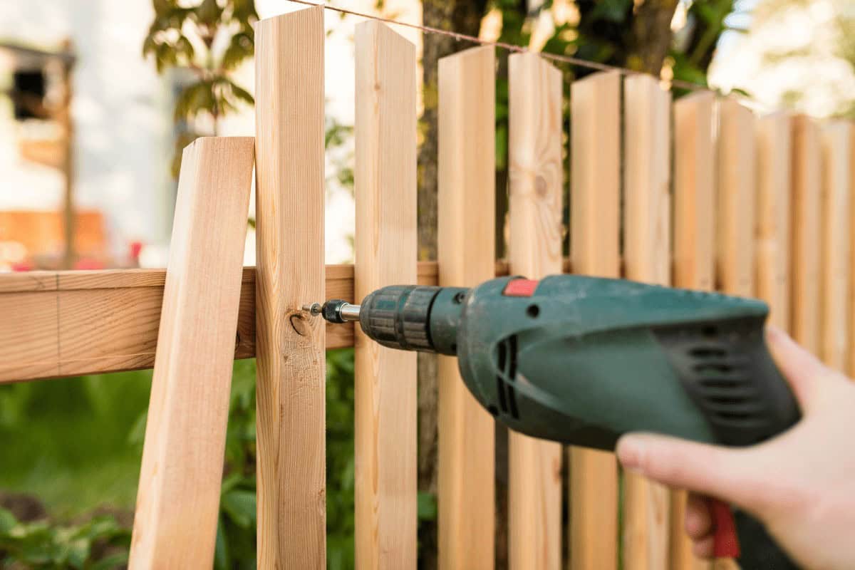A person uses a green electric drill to fasten wooden planks to form a fence. Sunlight shines on the scene, with blurred greenery in the background. The planks are evenly spaced and positioned vertically.