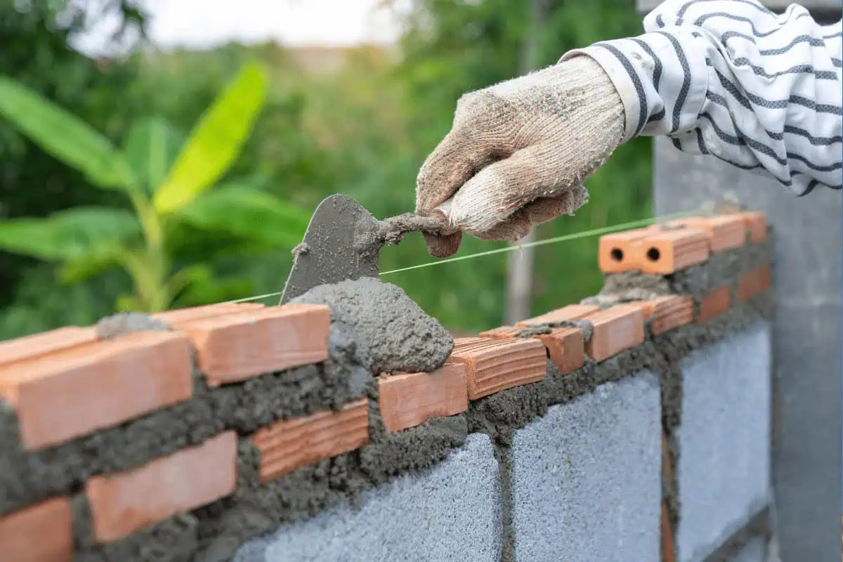 A person wearing a striped shirt and glove is using a trowel to lay and level mortar on a brick wall. The background includes green foliage and blurred plants.