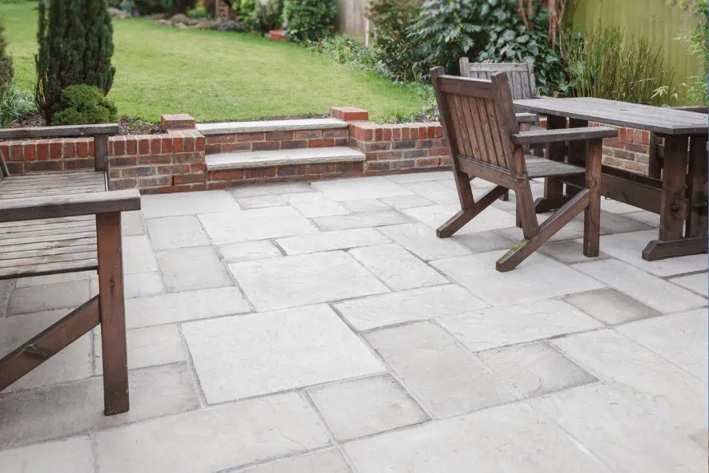 A backyard patio with light stone tiles, wooden chairs, and a table. The patio is bordered by red brick steps leading to a green lawn with shrubs and plants along the fence.