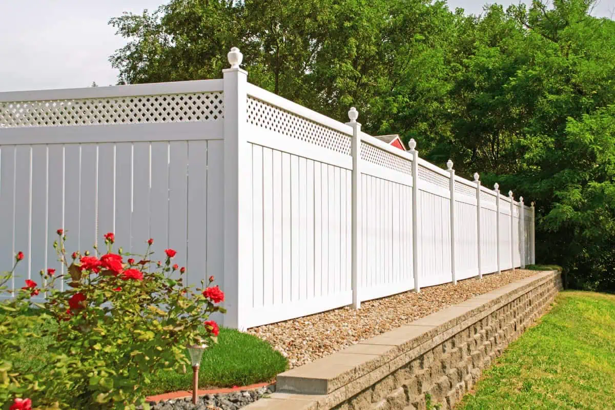 A tall white vinyl fence with lattice detailing on top runs along a stone retaining wall. Red flowers and green shrubs are in the foreground, with lush trees in the background under a clear sky.