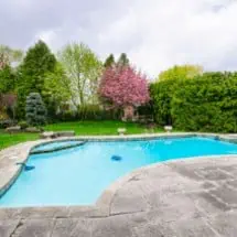 A backyard swimming pool with clear blue water rests beside a sleek pool deck, surrounded by a lush landscaped garden. Trees with pink blossoms and verdant shrubs create a serene backdrop. The sky is cloudy, and patio furniture is partially visible in the background.