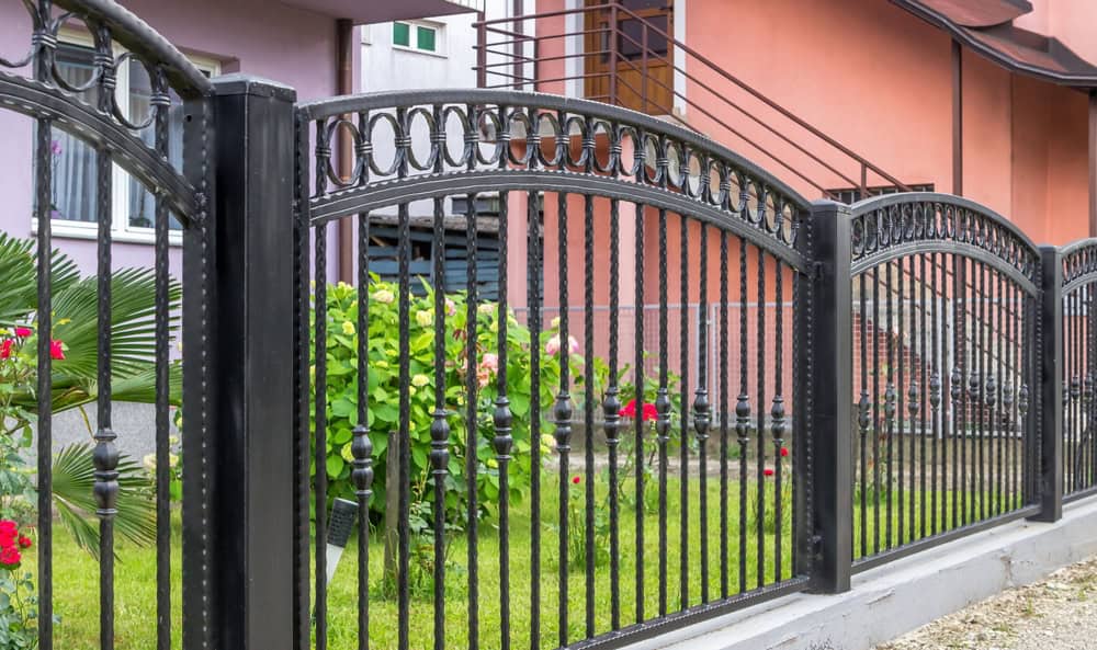 A decorative black aluminum fence with ornamental arches and circular patterns stands in front of a house. Behind the fence, lush greenery and blooming flowers abound. In the background, a pink and purple building is visible.
