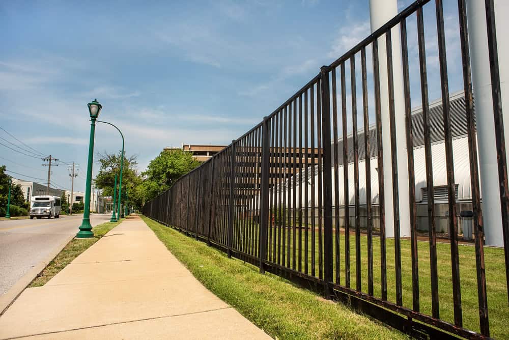A sidewalk runs alongside an aluminum fence and a grassy area. A green street lamp is visible on the left, and a large building with white panels is on the right. A truck is driving on the road under a clear blue sky.