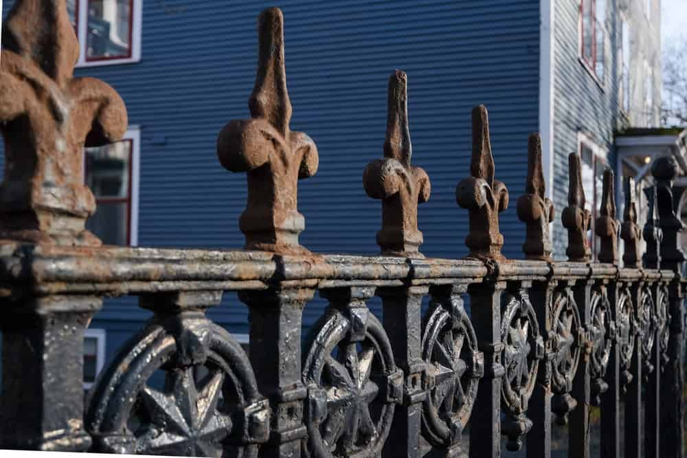 Close-up view of an ornate wrought iron fence with fleur-de-lis finials, partially rusted. An aluminum fencing section complements it nearby. A blue wooden building with white and red-trimmed windows stands in the background under a clear blue sky.