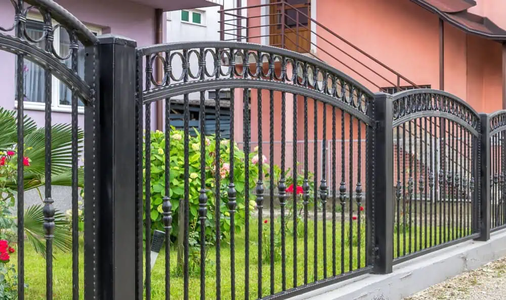 A decorative black aluminum fence with ornamental arches and circular patterns stands in front of a house. Behind the fence, lush greenery and blooming flowers abound. In the background, a pink and purple building is visible.