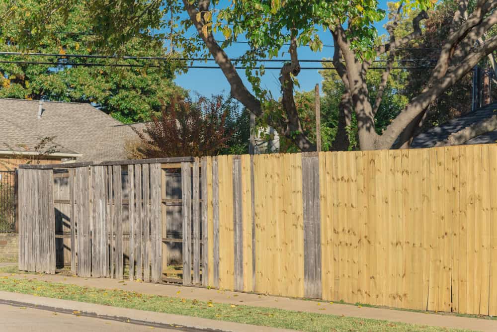 A wooden fence with sections of old, weathered gray wood beside newly installed, light-colored wood panels, standing along a suburban street with trees and houses in the background.