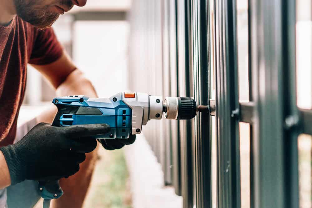 A person wearing black gloves uses a blue electric drill to fasten a screw into a metal fence or gate. The focus is on the hands and tool, with the background blurred.