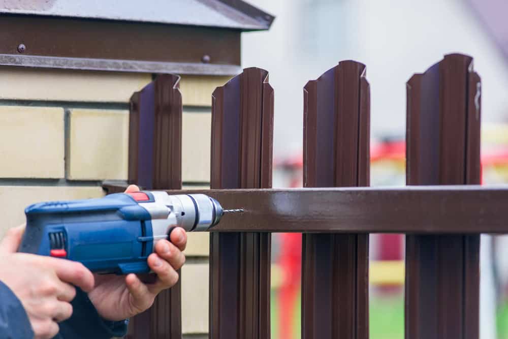 A person using a blue electric drill to make a hole in a brown metal fence, next to a beige brick pillar.
