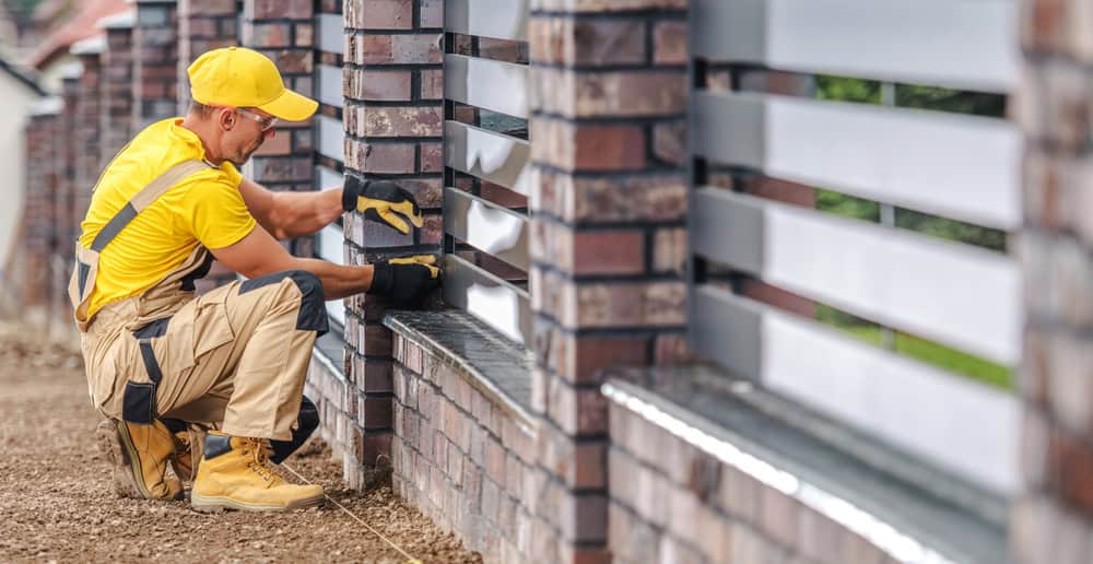 A construction worker in yellow clothing and boots installs horizontal metal panels on a brick fence outdoors.