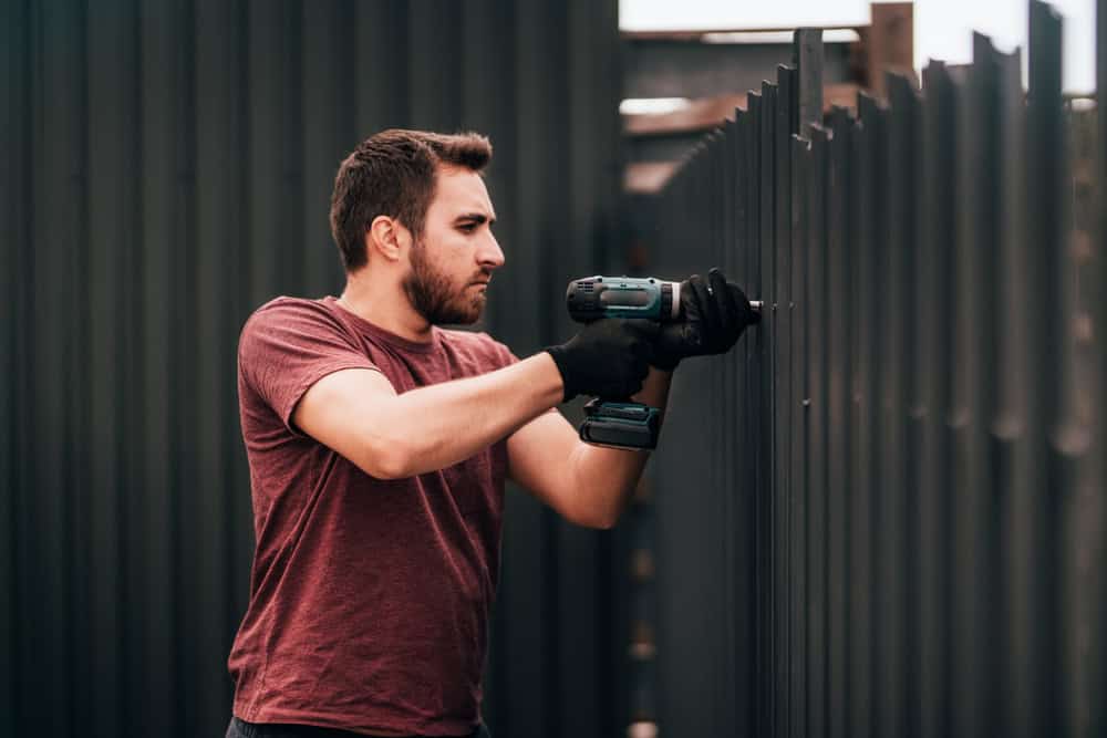 A man wearing a maroon t-shirt and black gloves uses a power drill to fasten a dark metal fence outdoors.