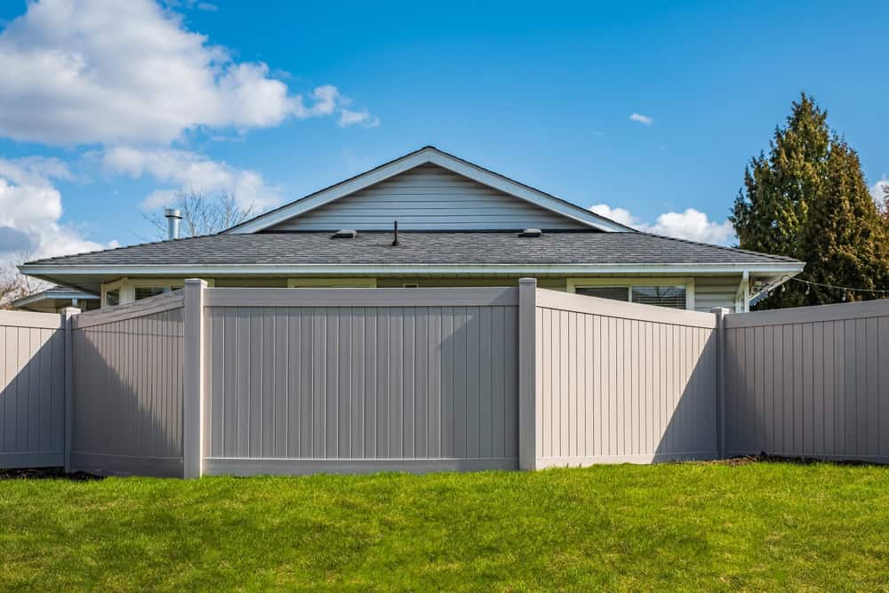 A gray vinyl privacy fence surrounds a green lawn in front of a single-story house with a gabled roof, under a blue sky with white clouds.