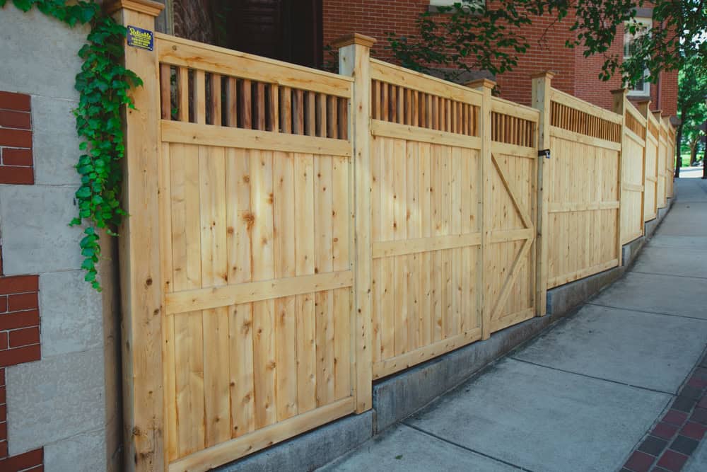 A tall wooden privacy fence with vertical slats on top runs along a sidewalk next to a brick building, with green ivy growing on the left side and trees providing partial shade overhead.