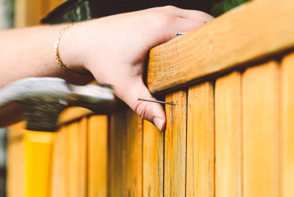Close-up of a hand holding a nail against a wooden fence while a hammer approaches. The person's wrist has a bracelet, and the wood shows a natural grain with a warm, light brown color.
