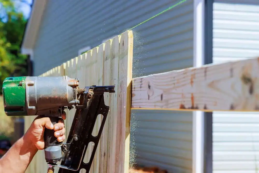 A person uses a nail gun to attach wooden planks to a fence. The wood is aligned with a green string for straightness. A house with light blue siding is visible in the background, and sawdust is seen in the air.