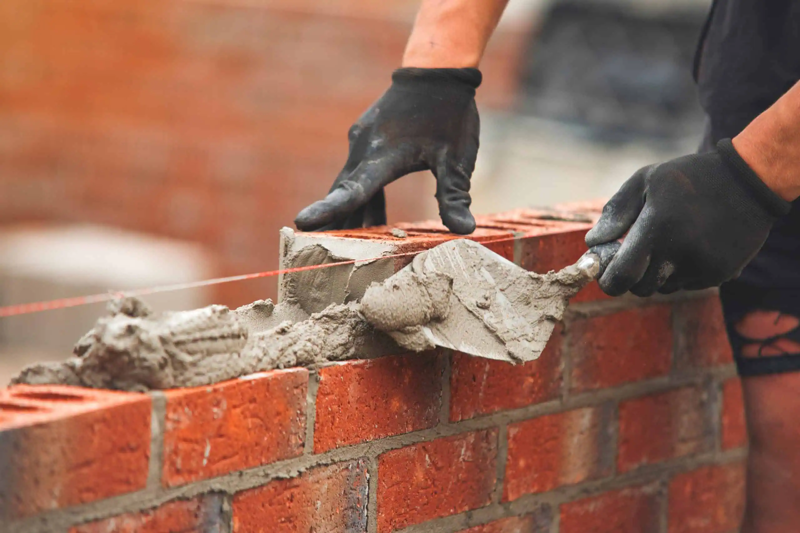 Close-up of a person wearing black gloves using a trowel to spread mortar on a brick wall under construction, aligning bricks with a guide string for accuracy.